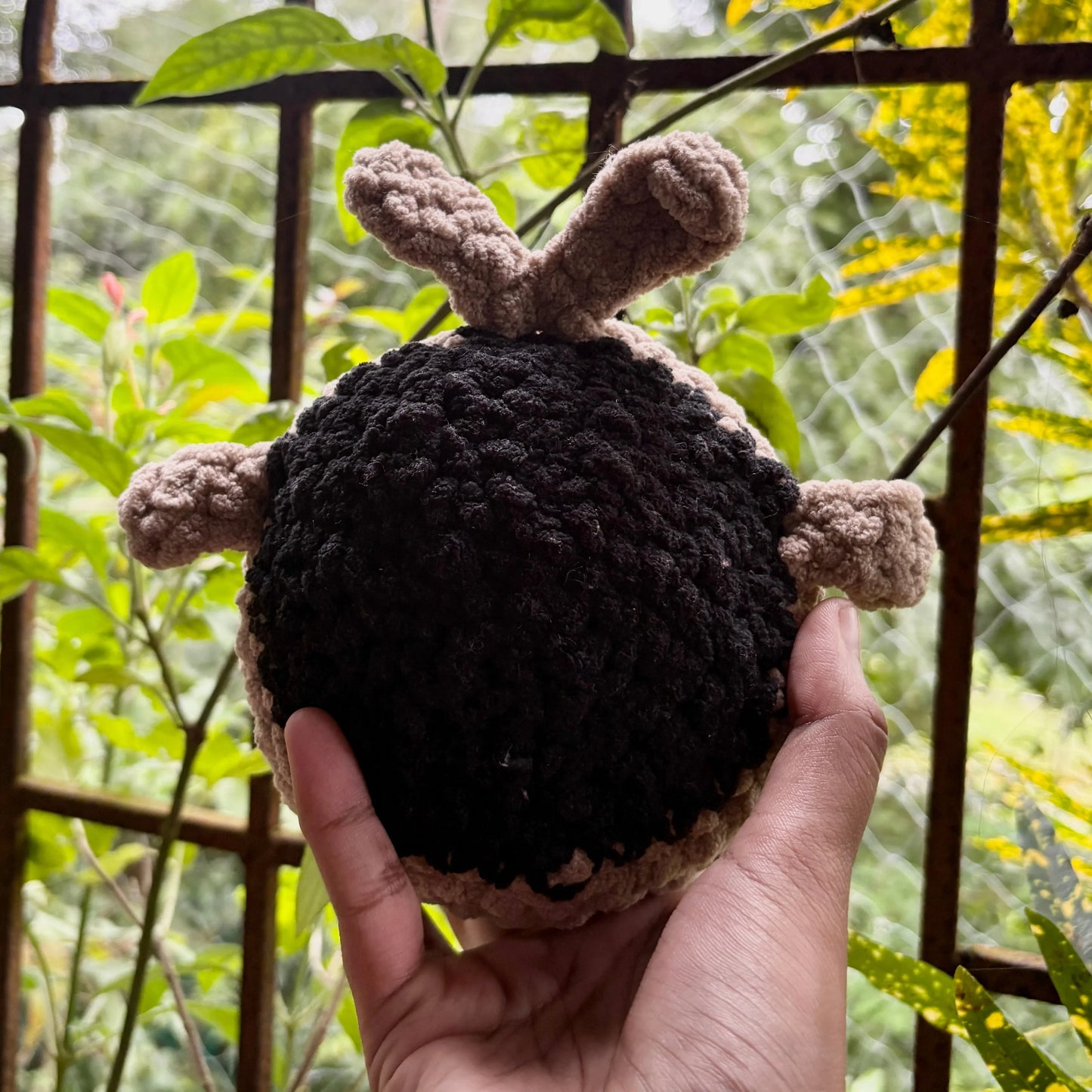 Hand holding a small crochet whale in front of a metal gate with greenery