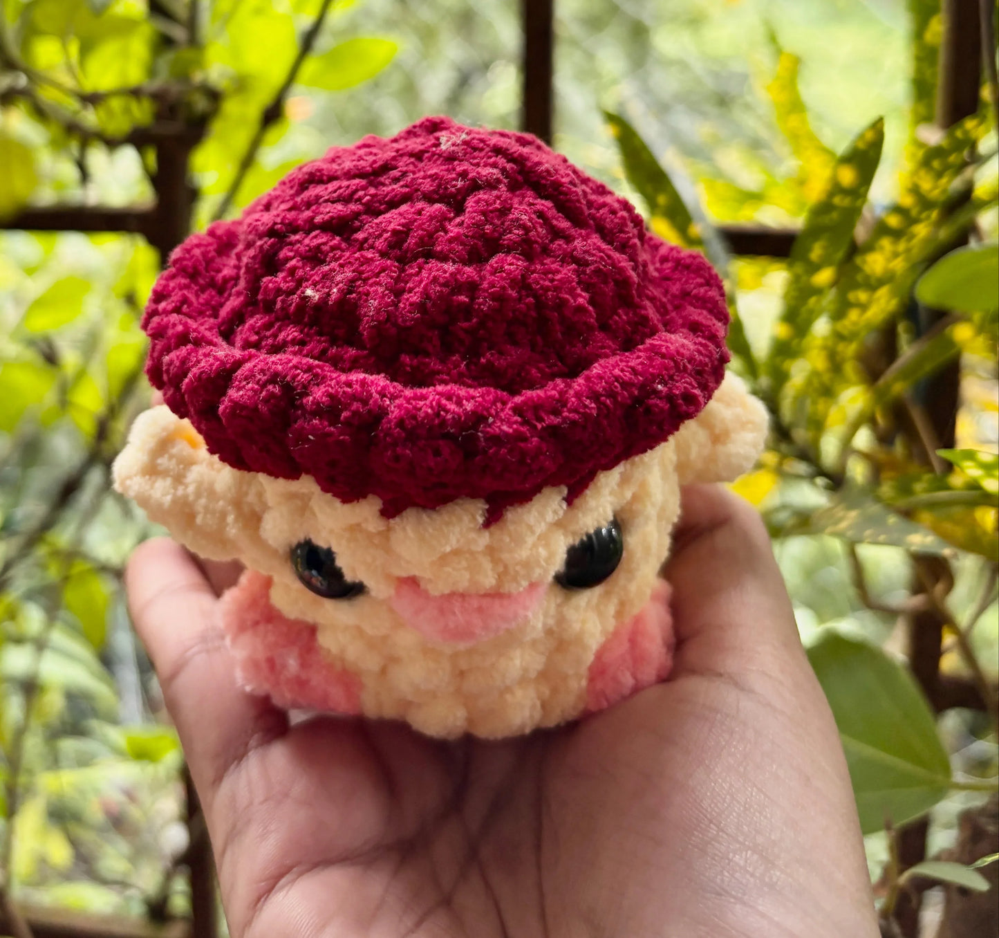 Hand holding a small plush toy with a red hat and black eyes against a blurred green background