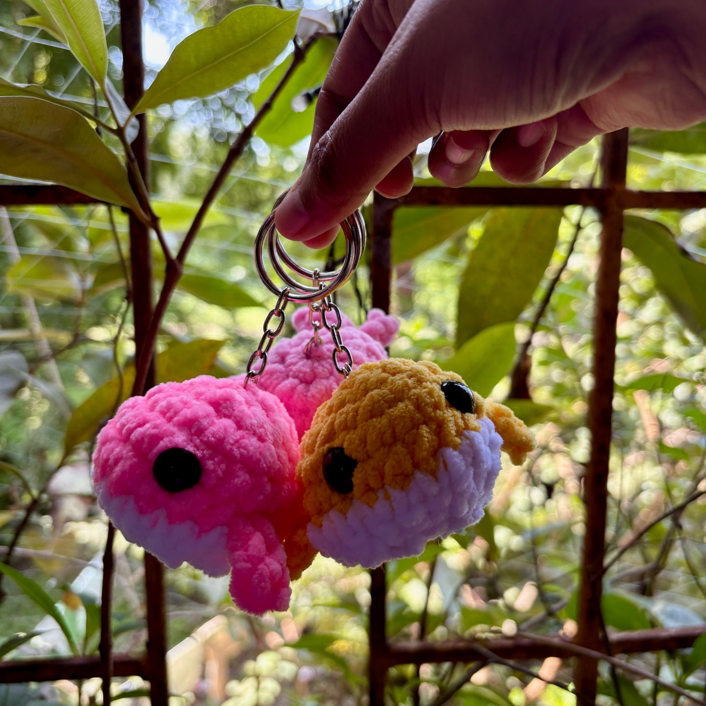Two colorful plush keychains hanging from a keyring, with a natural background of green leaves.