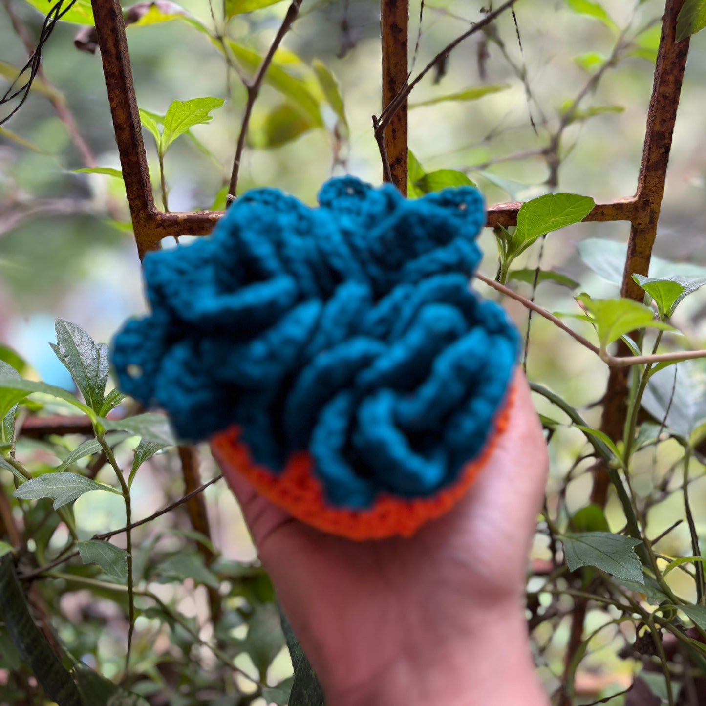 Hand holding a crochet Teal coaster set of 3 with orange pot against a blurred green outdoor background