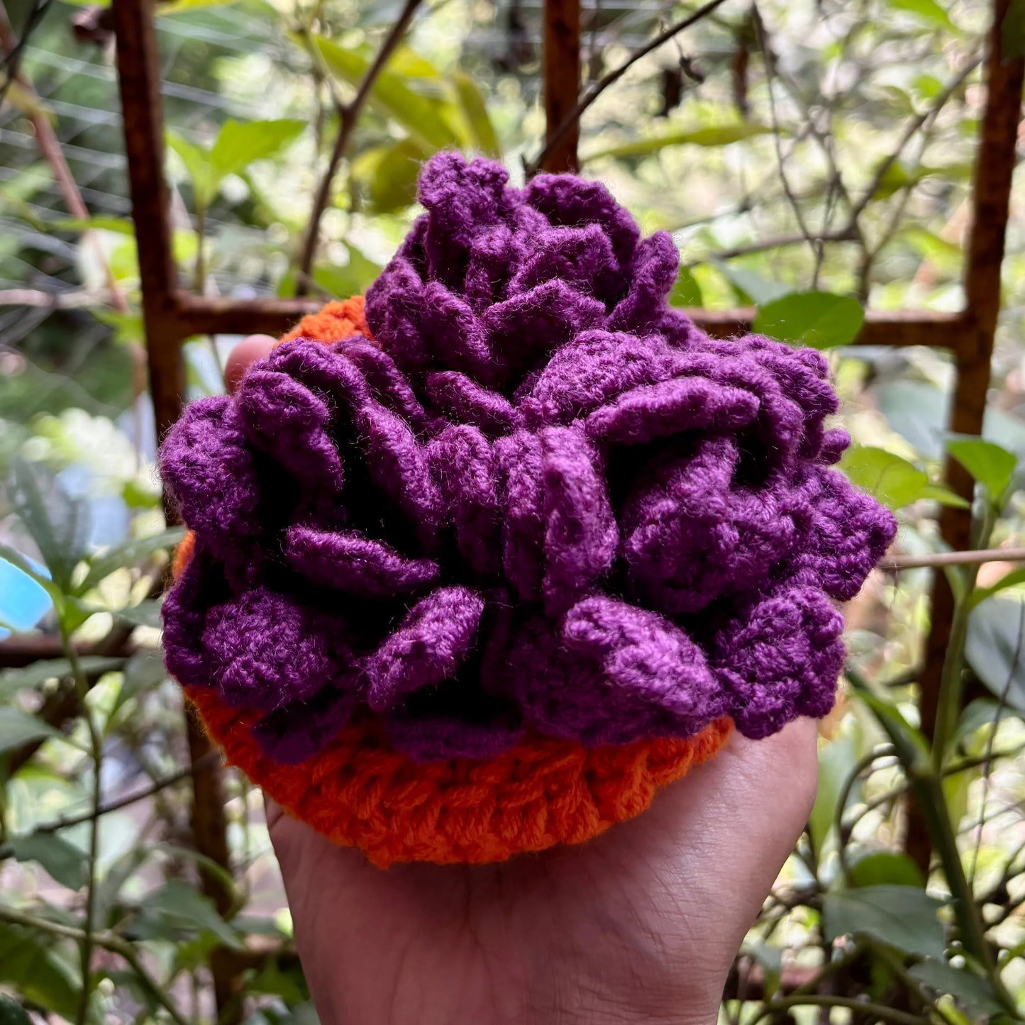 Hand holding a crochet purple coaster set of 3 with orange pot against a blurred green outdoor background