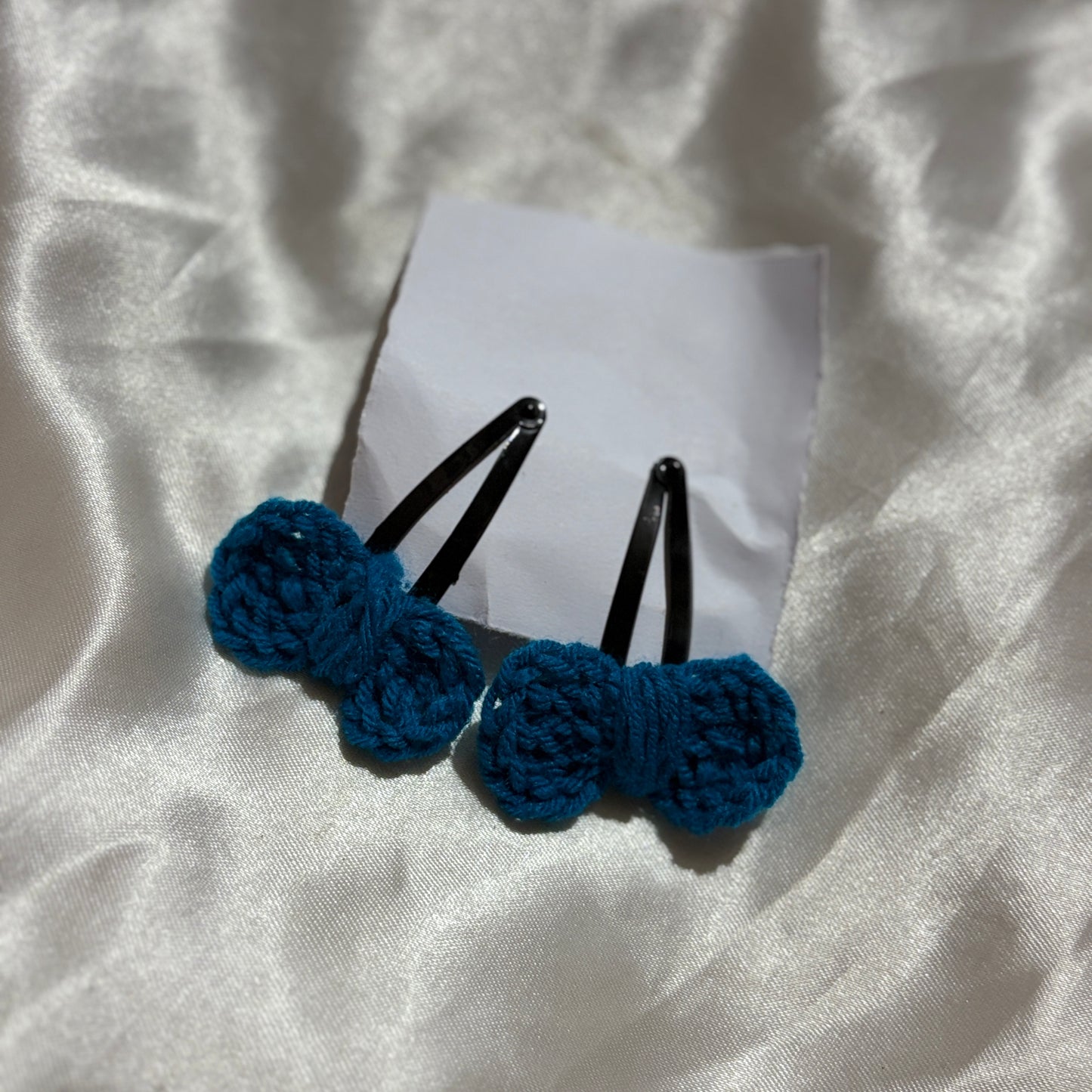Two blue hair bows with black clips on a white paper, placed on a textured white fabric background.