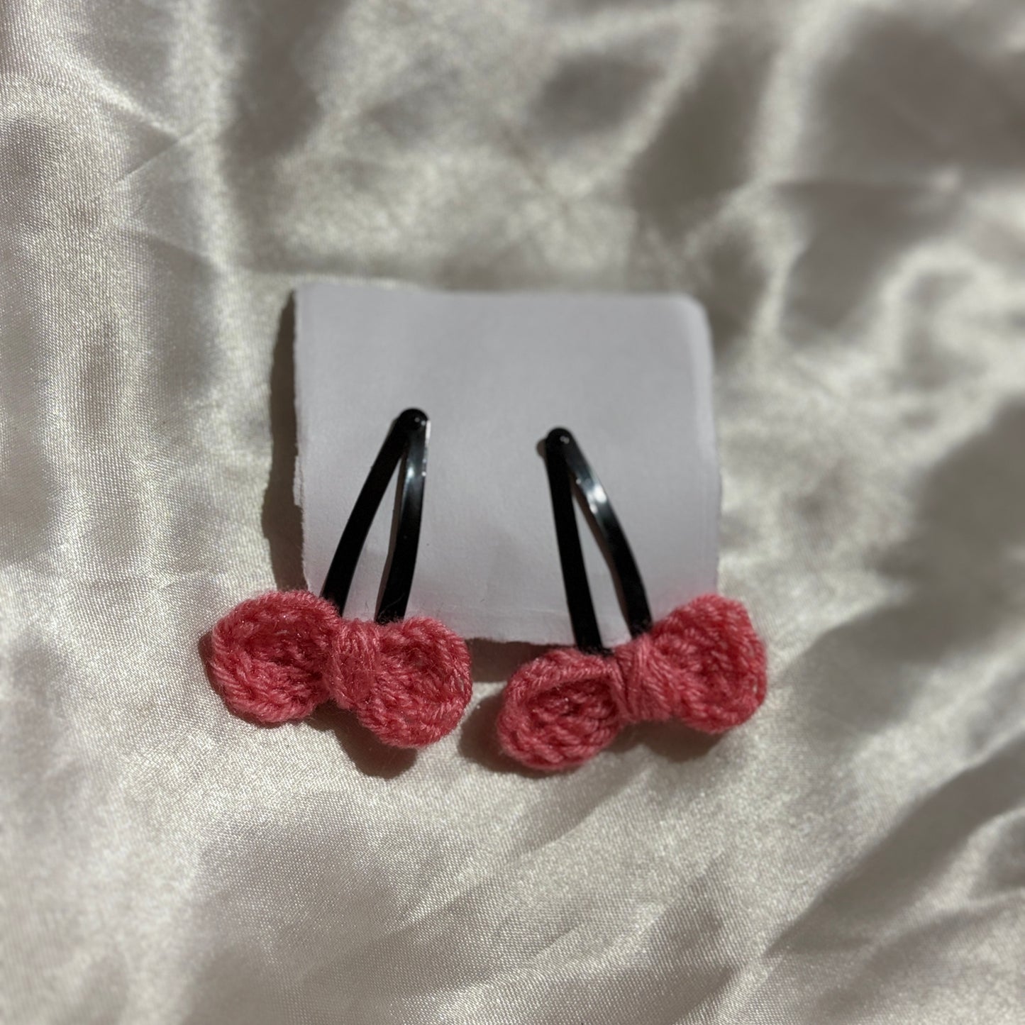 Pair of hair clips with red bow attachments on a white card, placed on a textured fabric background.