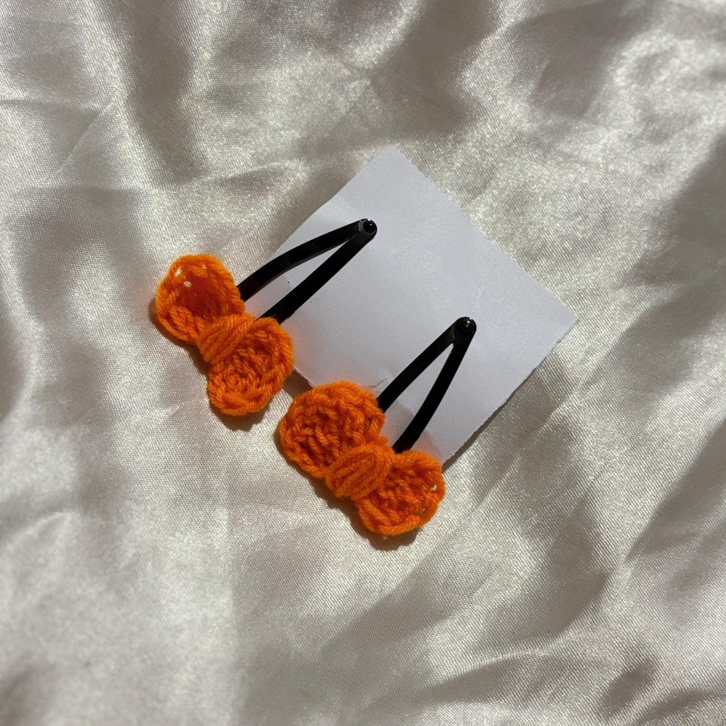 Two orange crocheted hair clips on a white card with a white fabric background