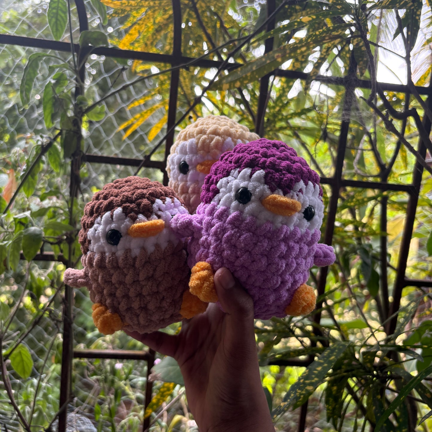 Three crocheted penguin toys held in front of a natural background with greenery.