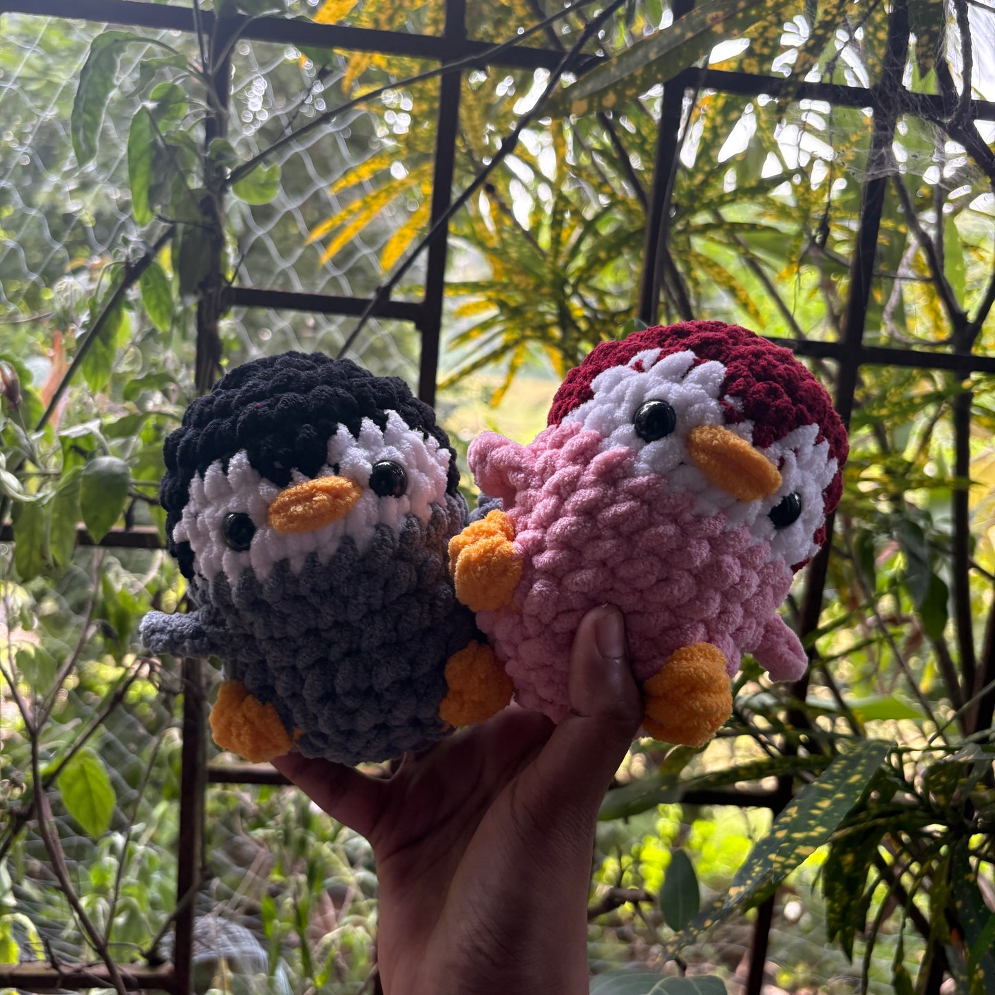 Two crocheted penguin toys held in front of a natural background with greenery.