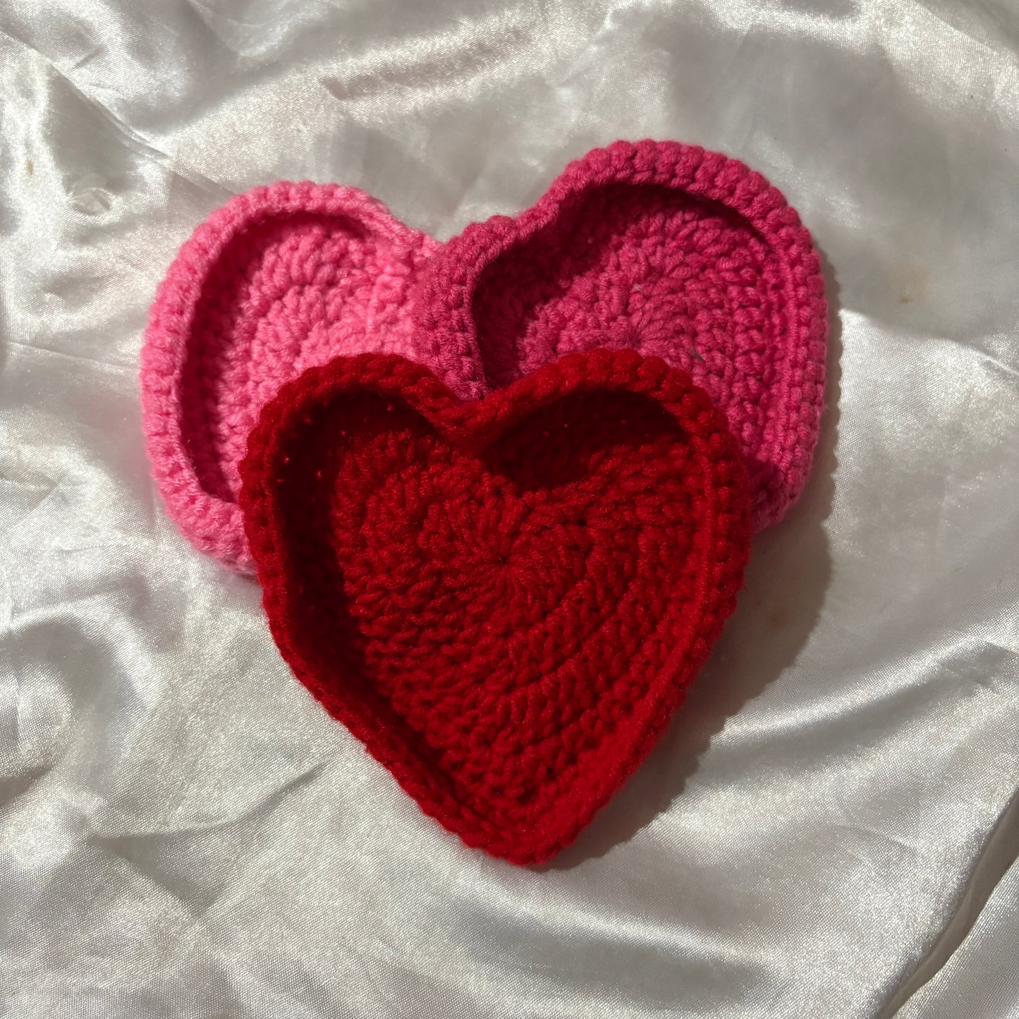 Two crocheted heart-shaped coasters, one red and one pink, on a white fabric background.