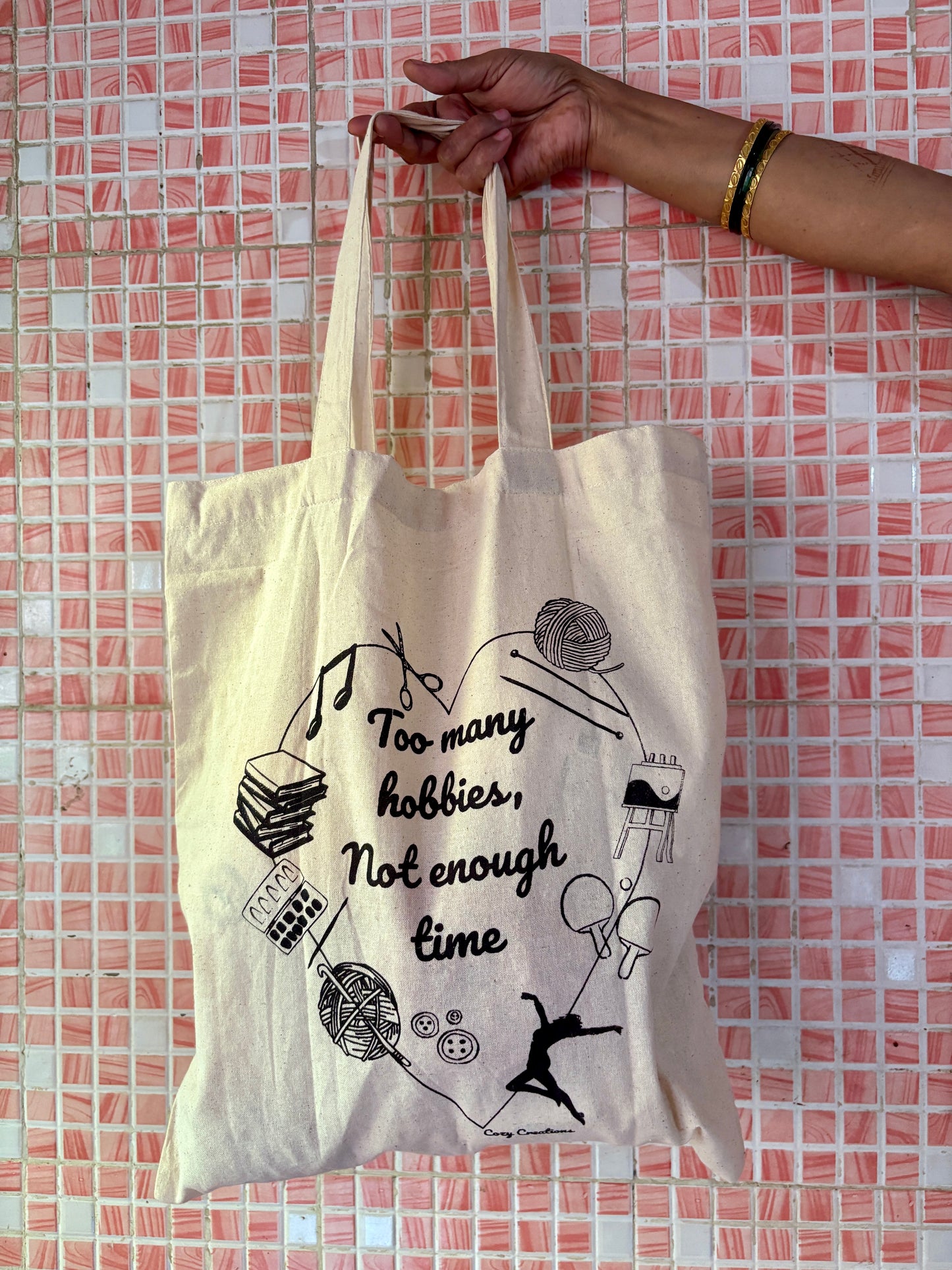 Tote bag with text and graphics held by a hand against a tiled wall background - too many hobbies not enough time