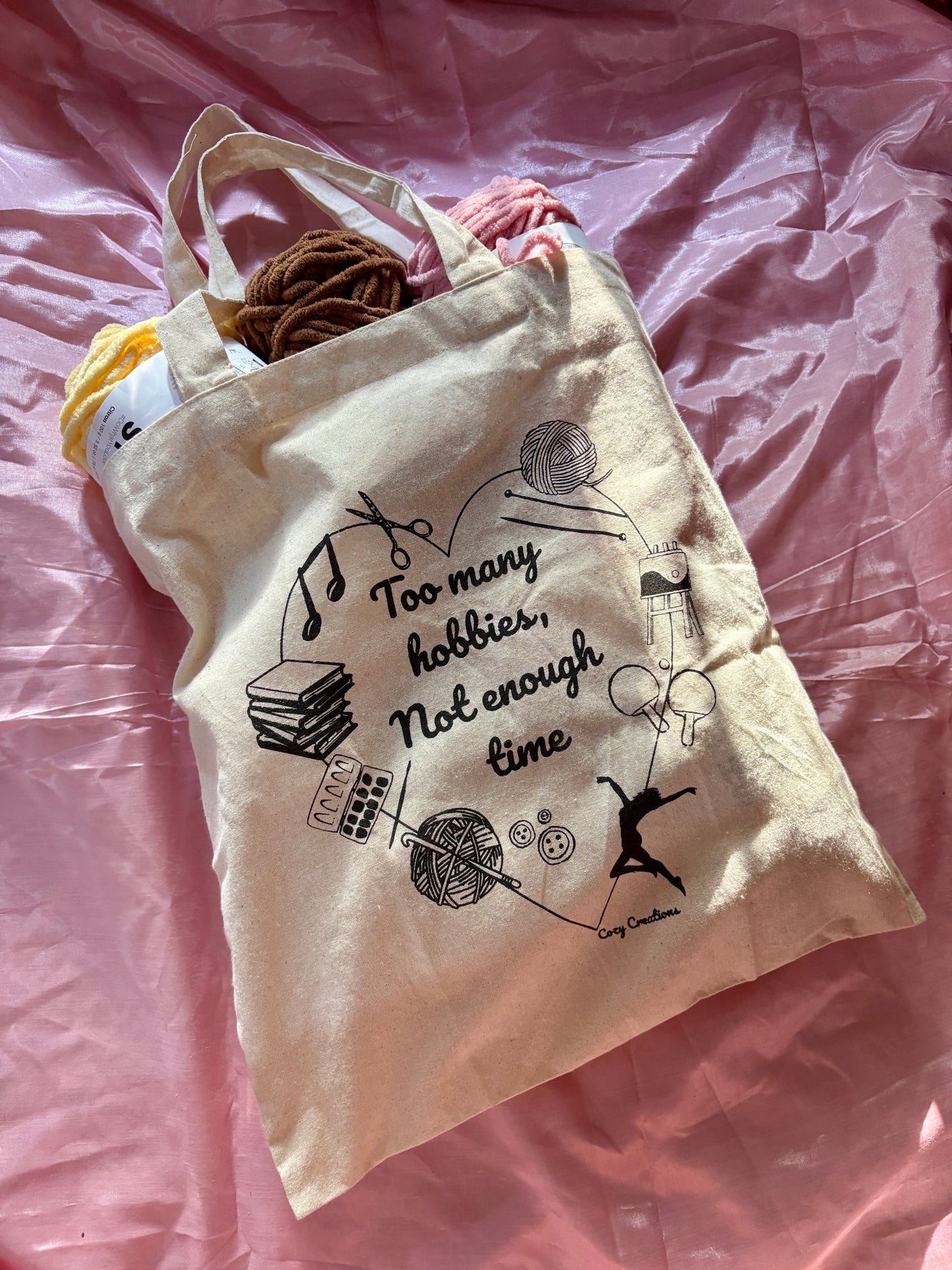 Tote bag with black graphics and text on a pink fabric background - too many hobbies not enough time