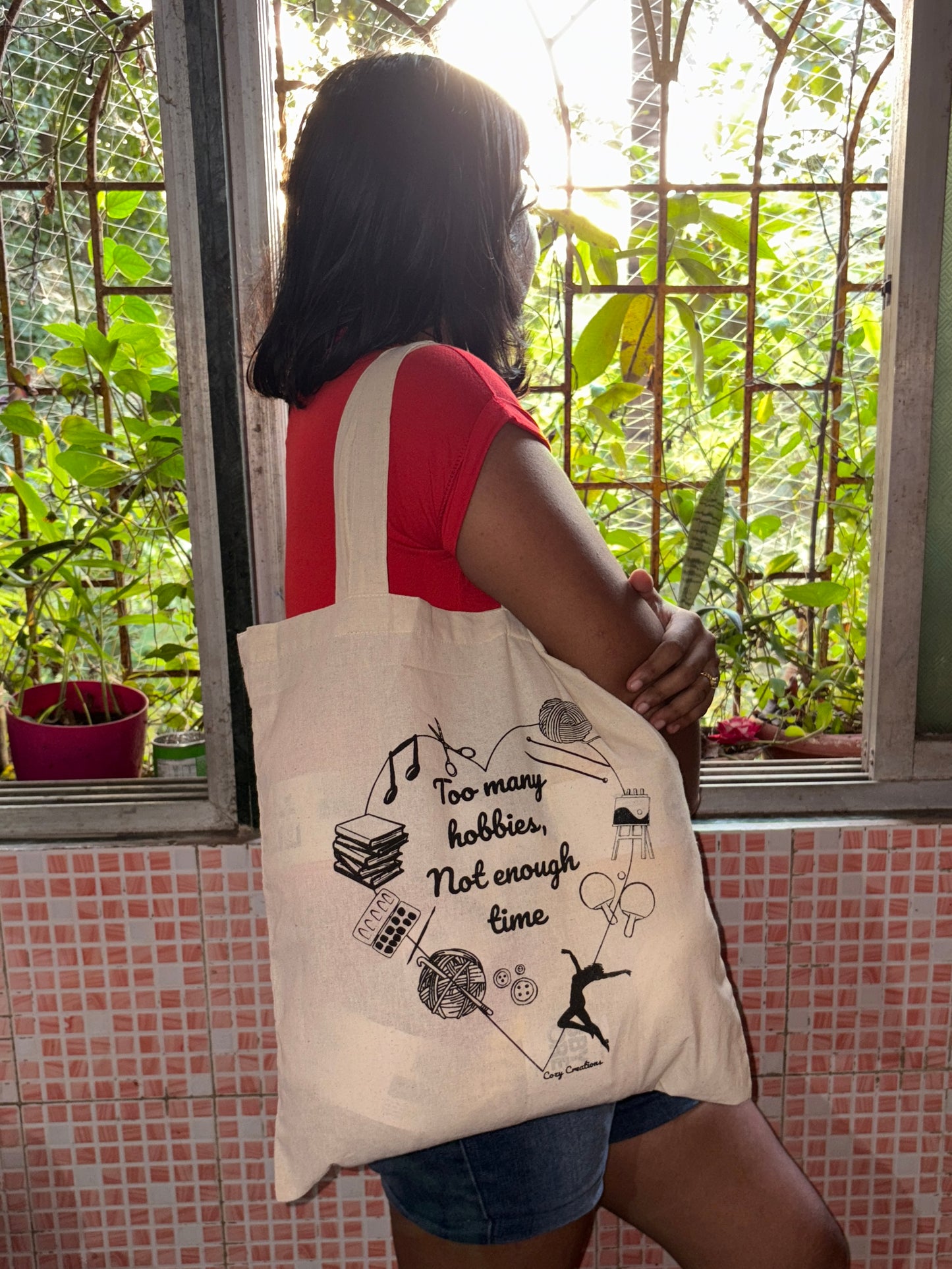 Person holding a tote bag with text and graphics in front of a window with plants. too many hobbies not enough time