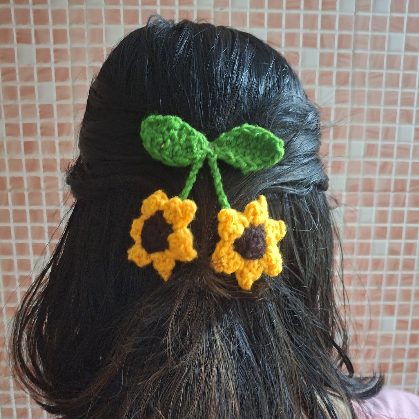 Person with black hair wearing a green and yellow crocheted hair flower accessory against a tiled wall.