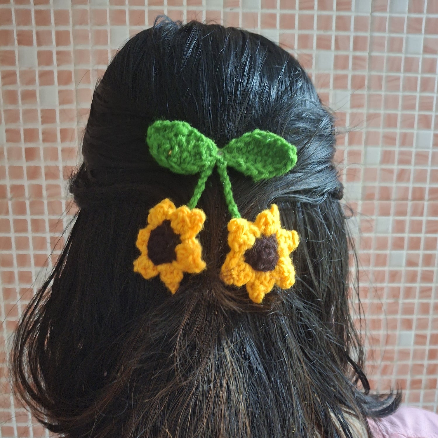 Person with black hair wearing a green and yellow crocheted hair flower accessory against a tiled wall.