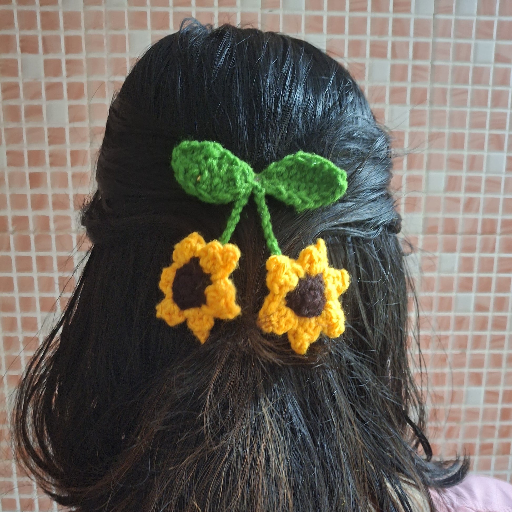 Person with black hair wearing a green and yellow crocheted hair flower accessory against a tiled wall.