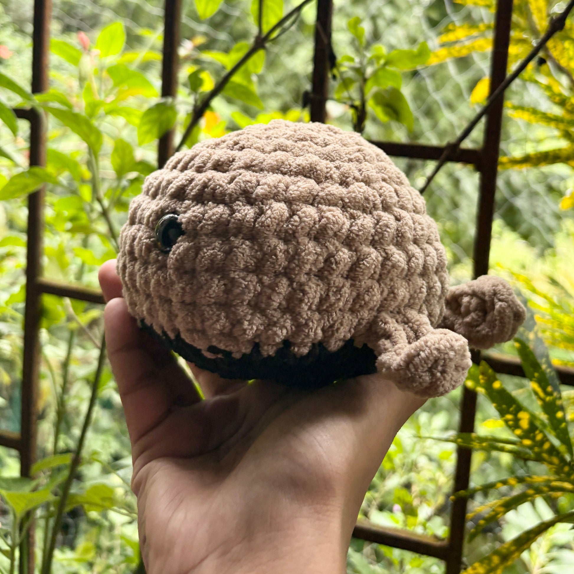 Hand holding a small crochet whale in front of a metal gate with greenery
