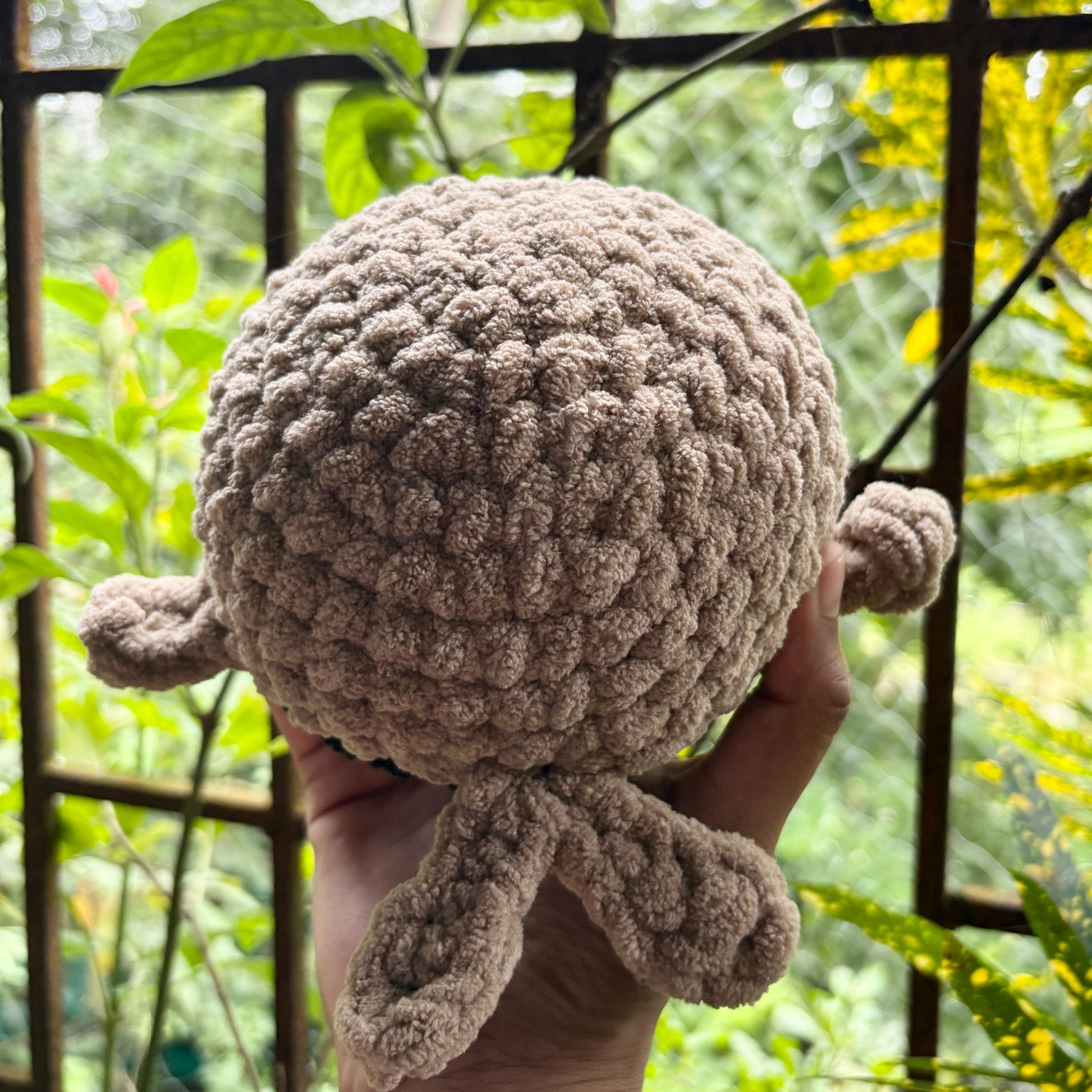 Hand holding a small crochet whale in front of a metal gate with greenery