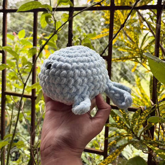 Hand holding a small crochet whale in front of a metal gate with greenery