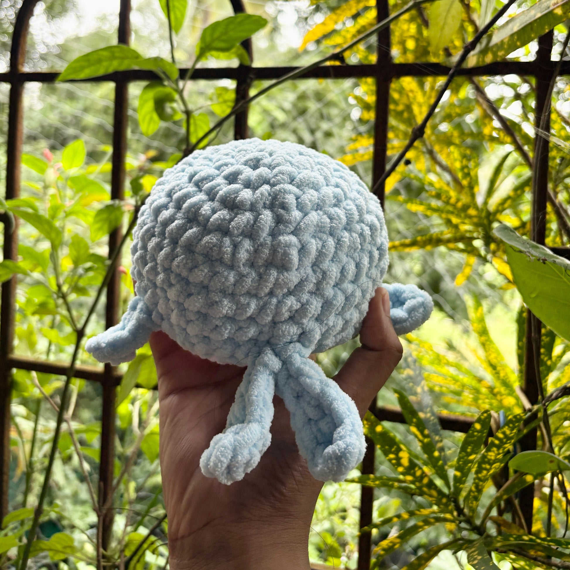 Hand holding a small crochet whale in front of a metal gate with greenery
