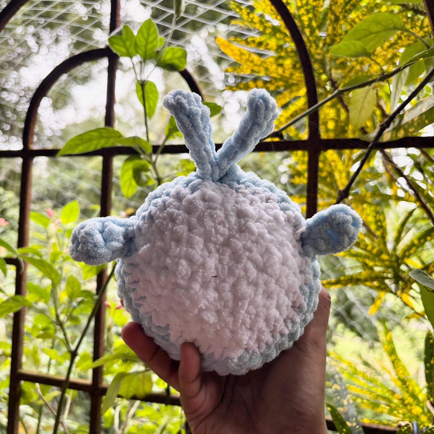 Hand holding a small crochet whale in front of a metal gate with greenery