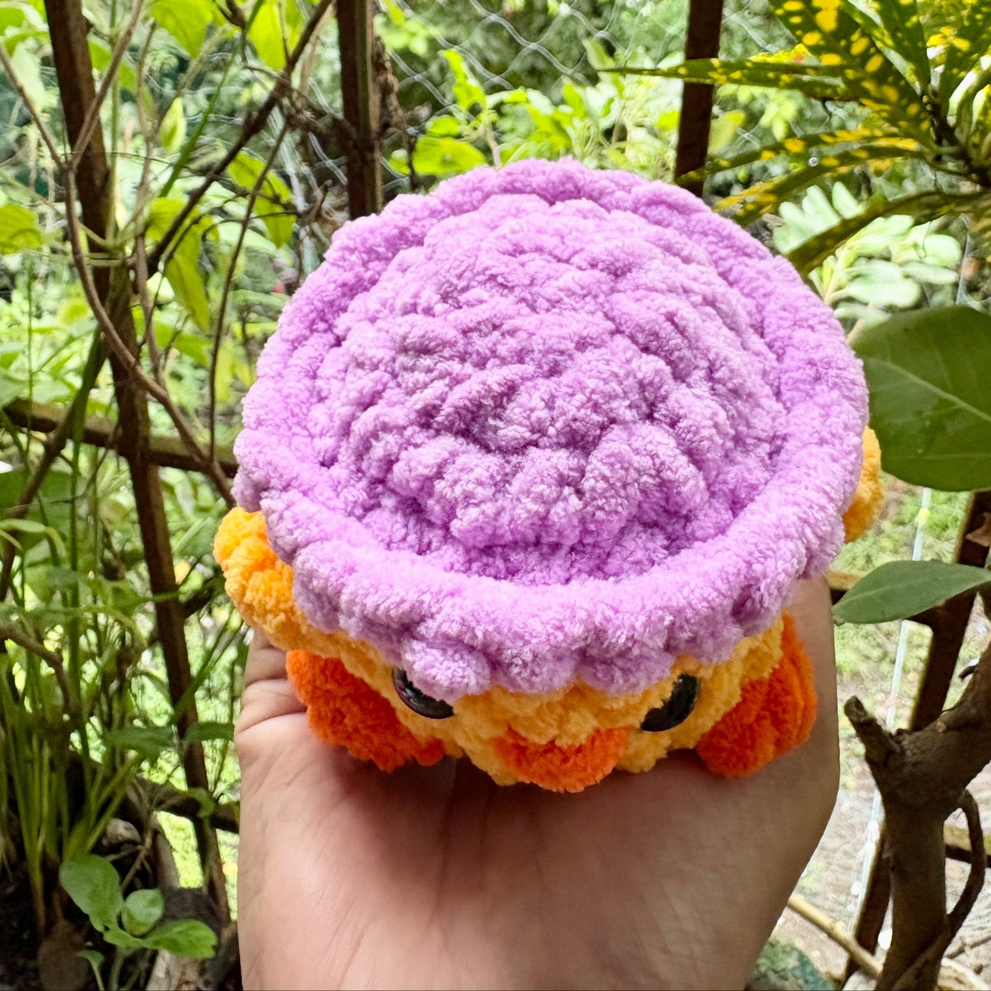 Hand holding a purple and orange plush toy against a natural background