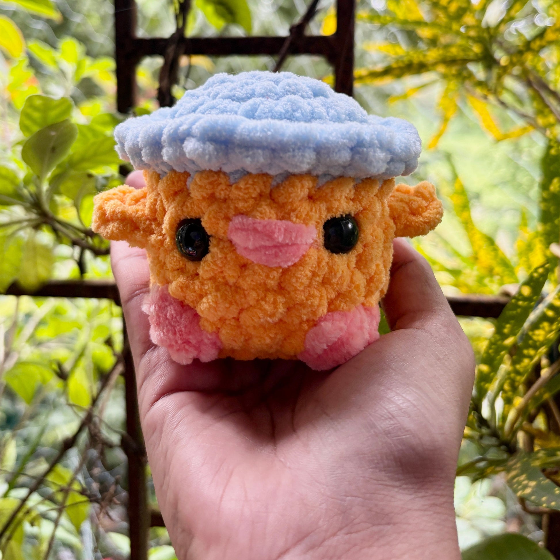 Hand holding a small orange plush toy with a blue hat against a blurred green background