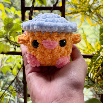 Hand holding a small orange plush toy with a blue hat against a blurred green background