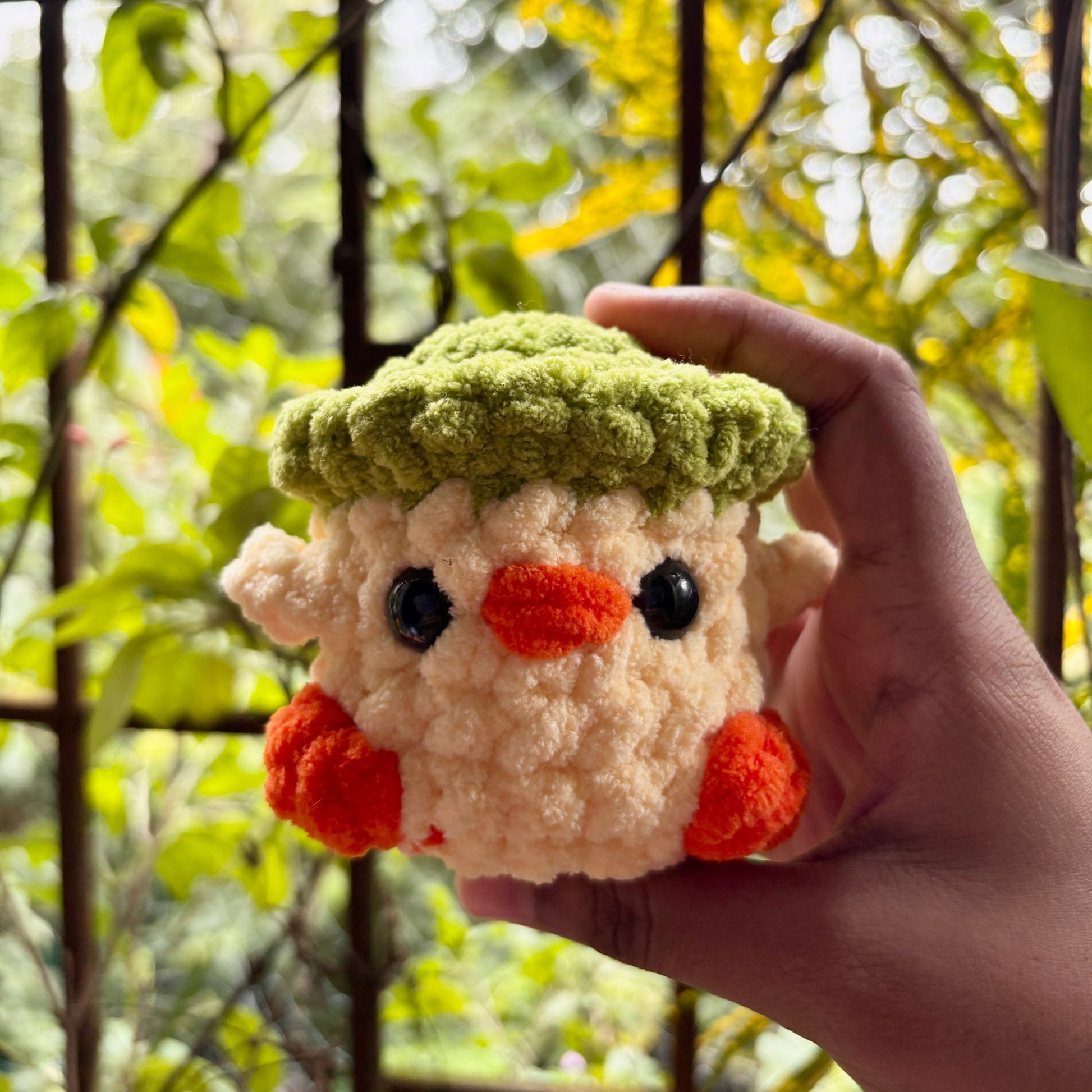 Hand holding a small crochet character with a green hat and orange feet against a blurred natural background