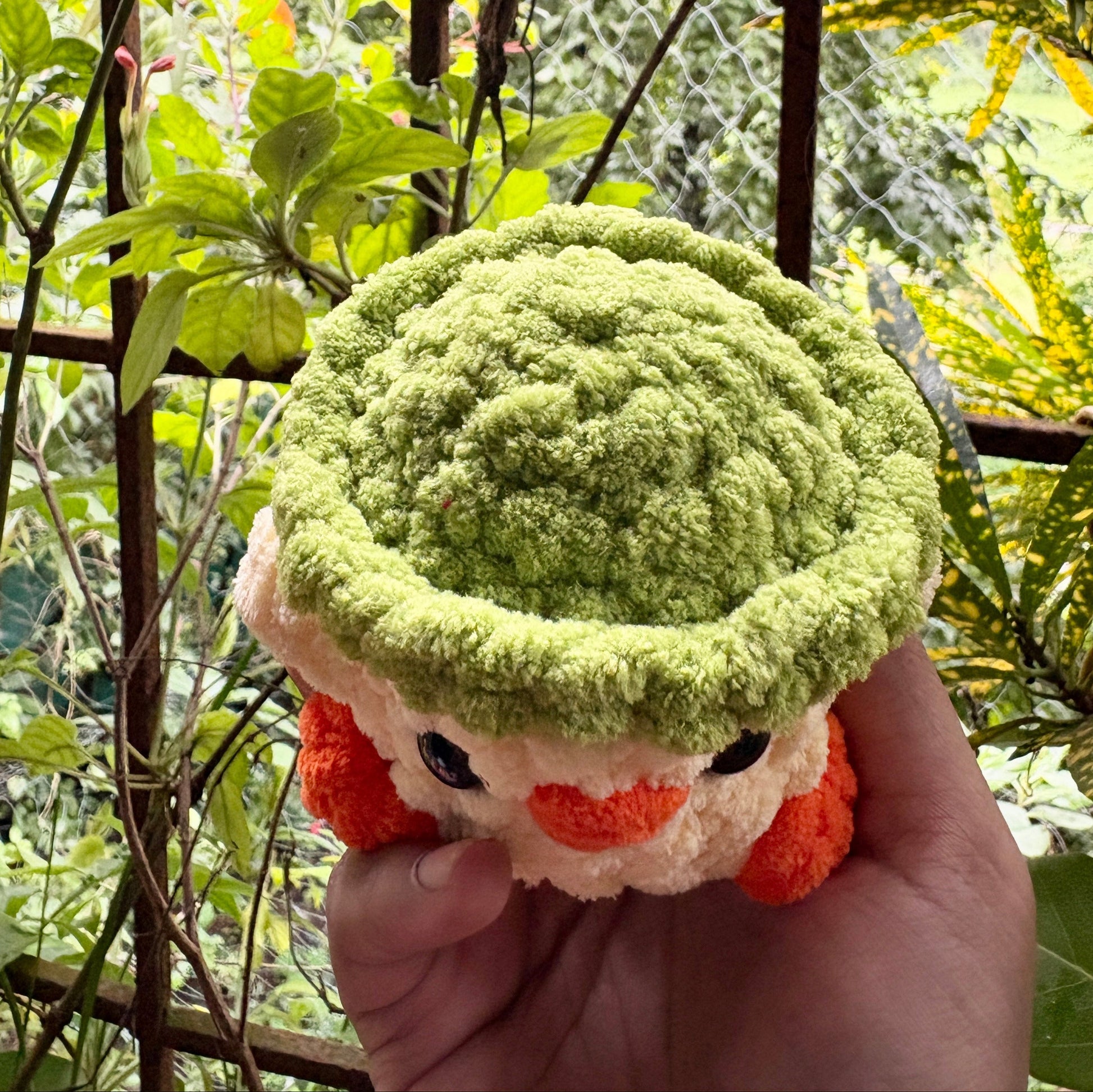 Plush toy resembling a character with a green hat and orange accents held in front of a natural background.
