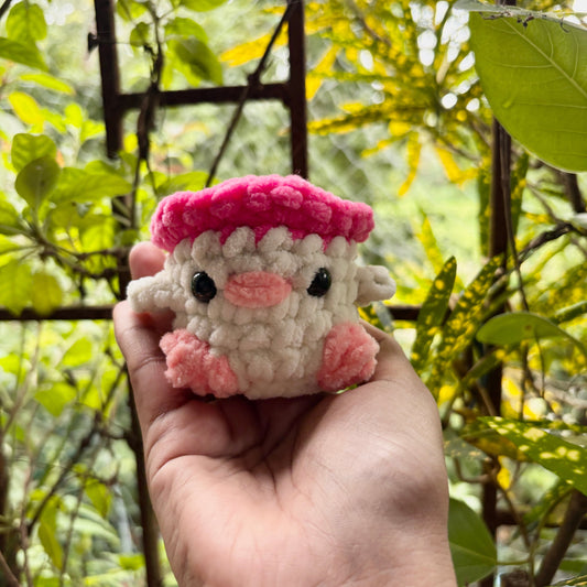 Hand holding a small crochet sheep toy with a pink hat against a natural background