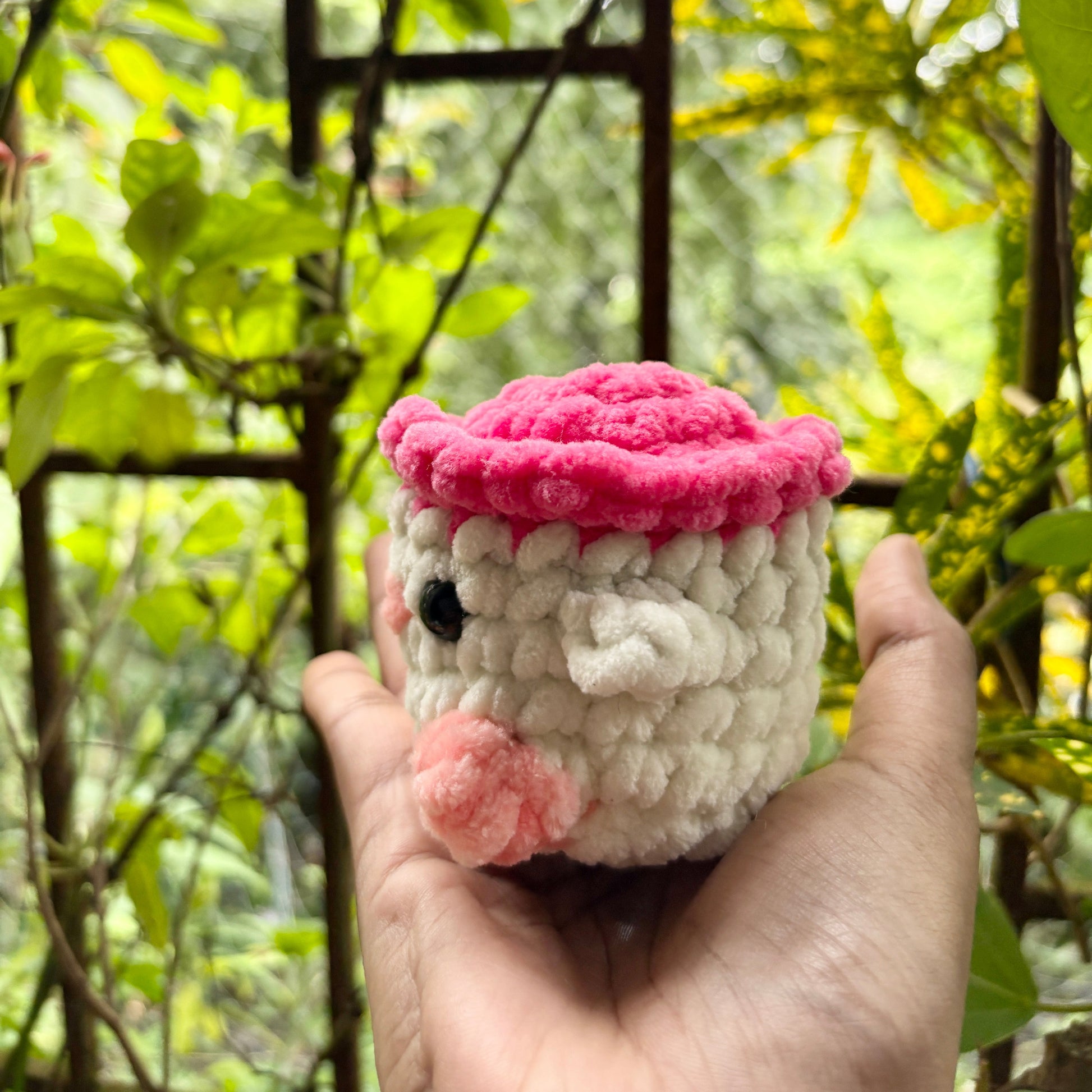 Hand holding a small knitted toy with a pink top against a blurred green outdoor background