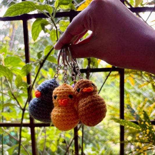 Three crocheted duck keychains held by a hand with a blurred green outdoor background
