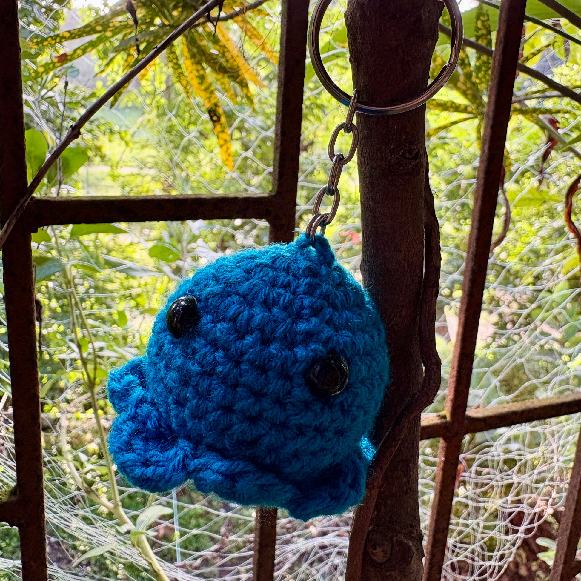 Blue crocheted keychain with black eyes hanging from a chain on a rusty metal structure with green foliage in the background.