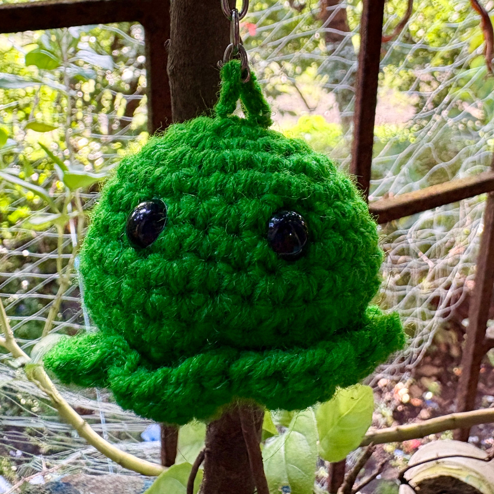 Green crocheted toy hanging from a tree branch with a blurred natural background