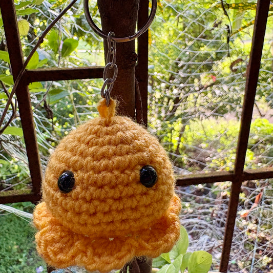 Crocheted keychain with a round, beige body and black eyes hanging outdoors.