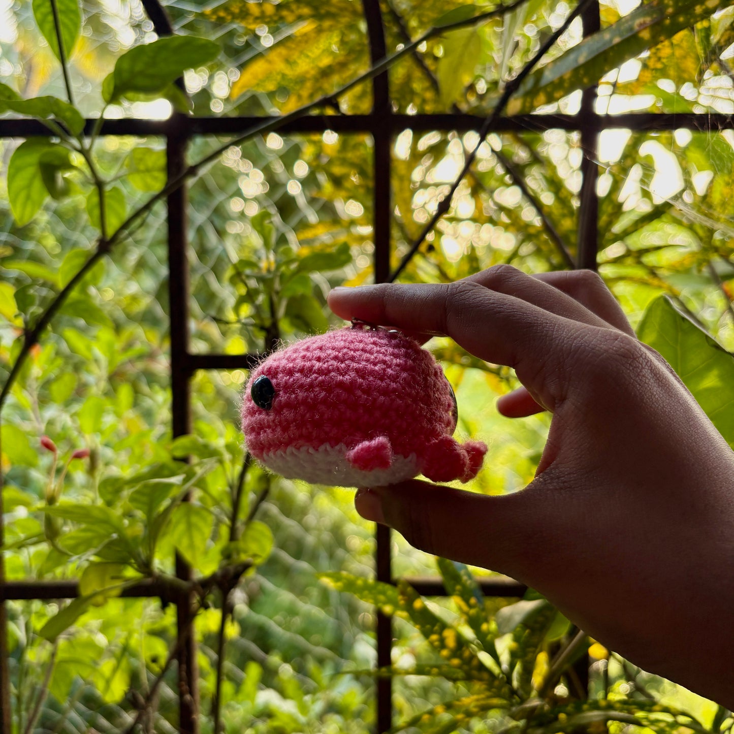 Hand holding a small yellow crocheted whale against a green leafy background