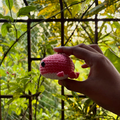 Hand holding a small yellow crocheted whale against a green leafy background
