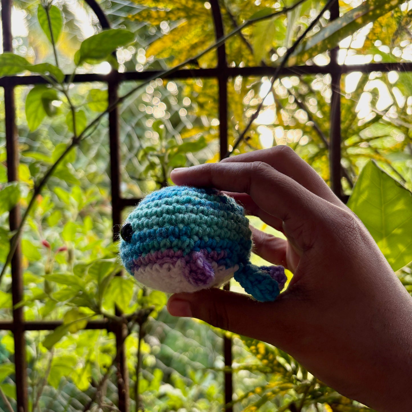Hand holding a small crocheted whale against a green leafy background