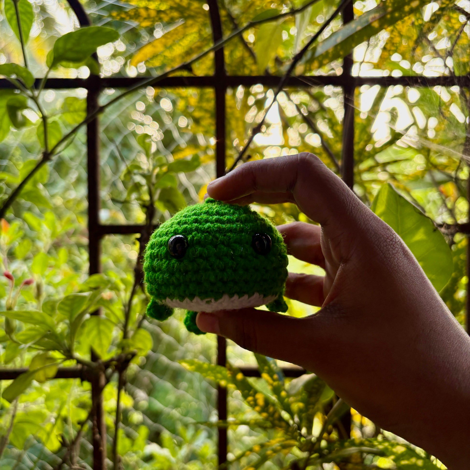 Hand holding a green crochet whale in front of a blurred natural background
