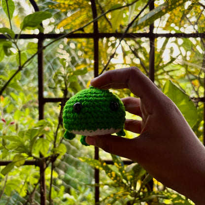 Hand holding a green crochet whale in front of a blurred natural background