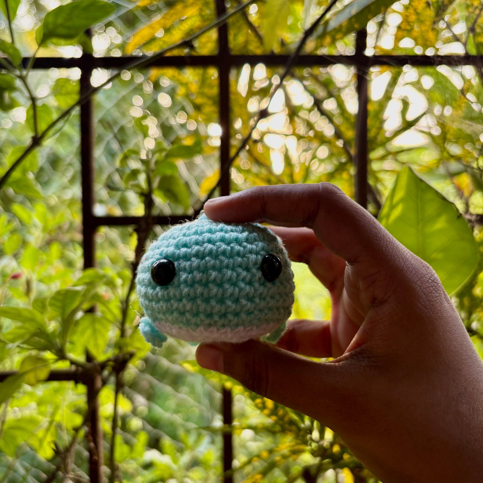 Hand holding a small blue crocheted whale against a blurred green leafy background