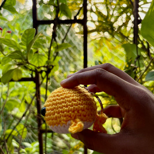 Hand holding a small yellow crocheted whale against a green leafy background