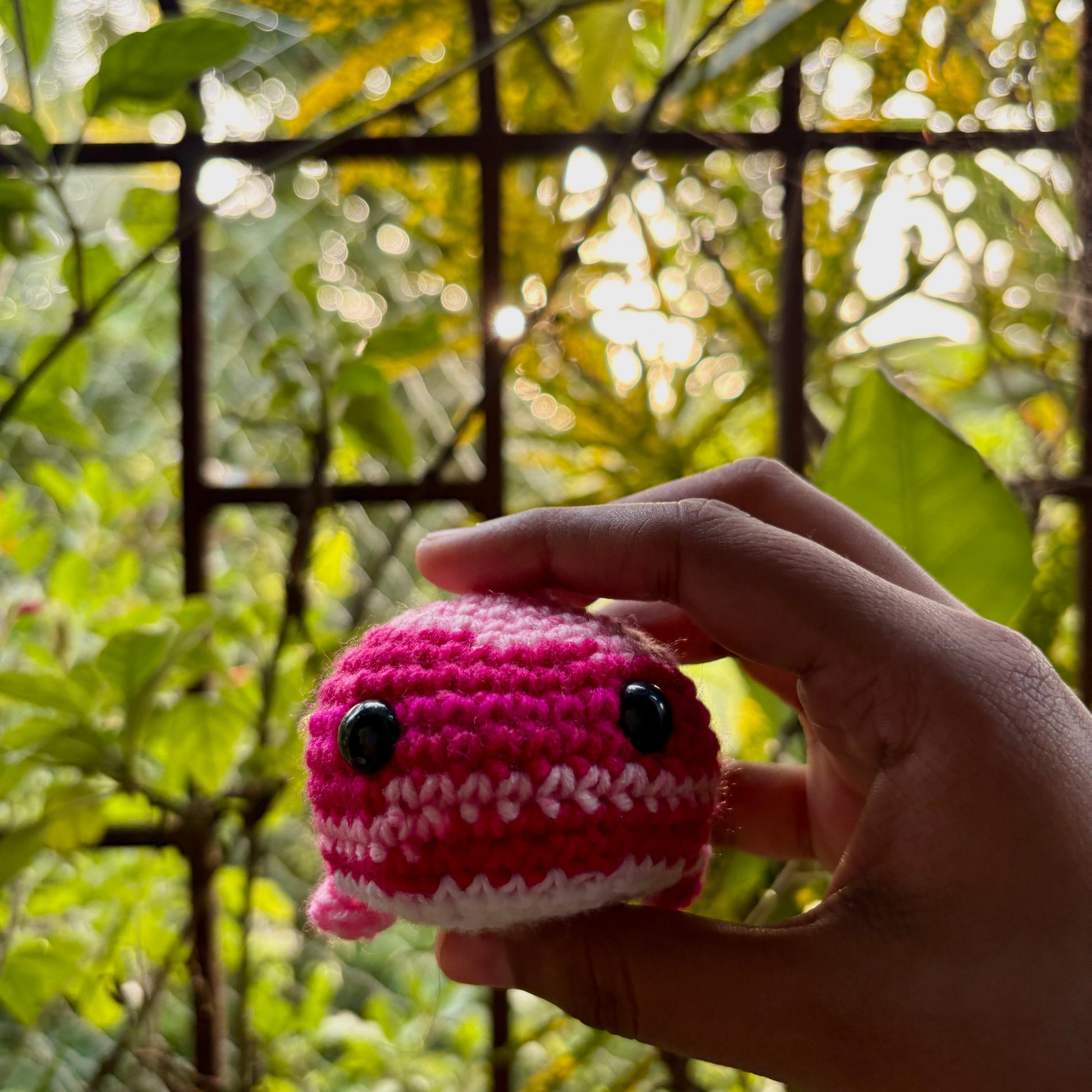 Hand holding a small yellow crocheted whale against a green leafy background
