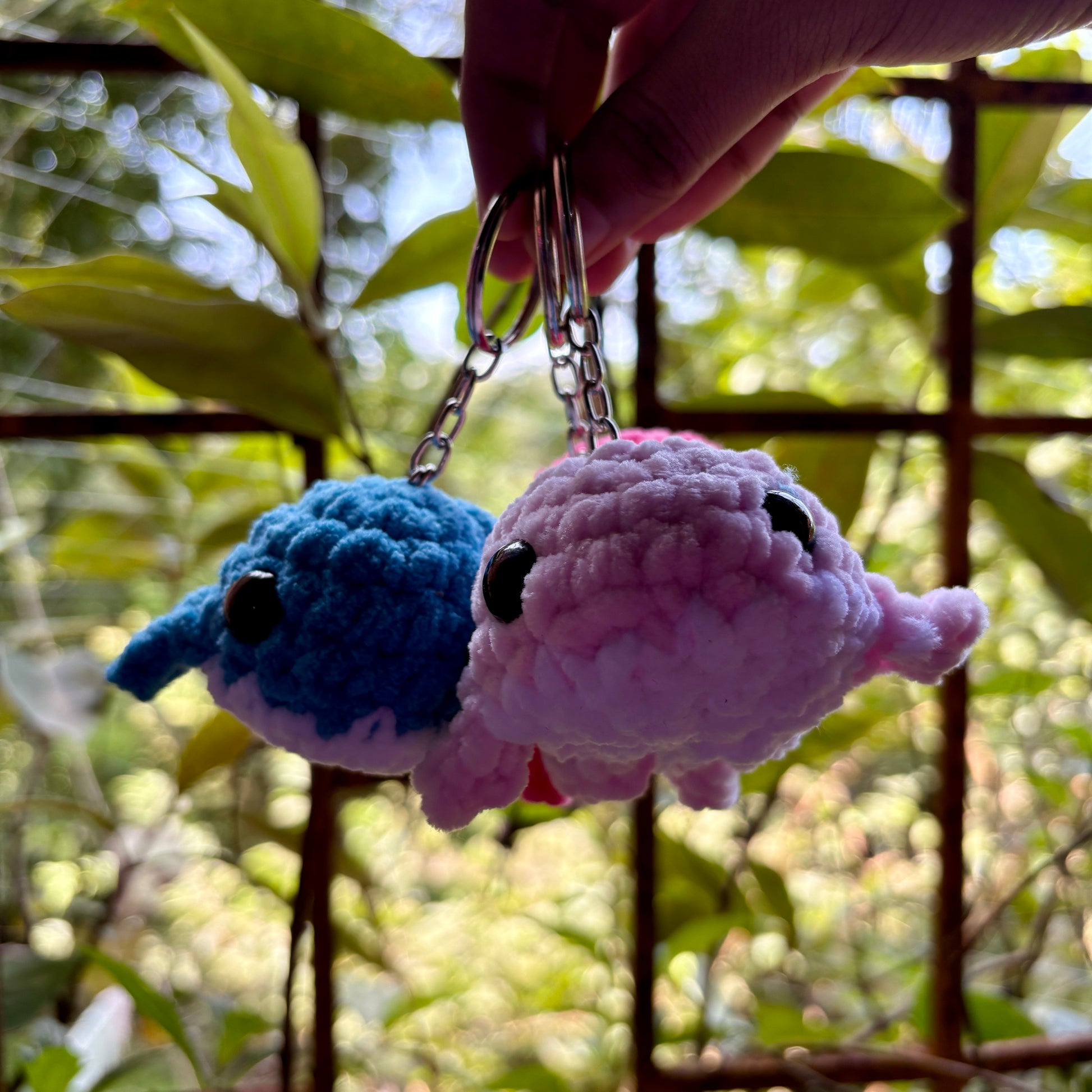 Two crochet whale keychains, one blue and one pink, held against a blurred green outdoor background.