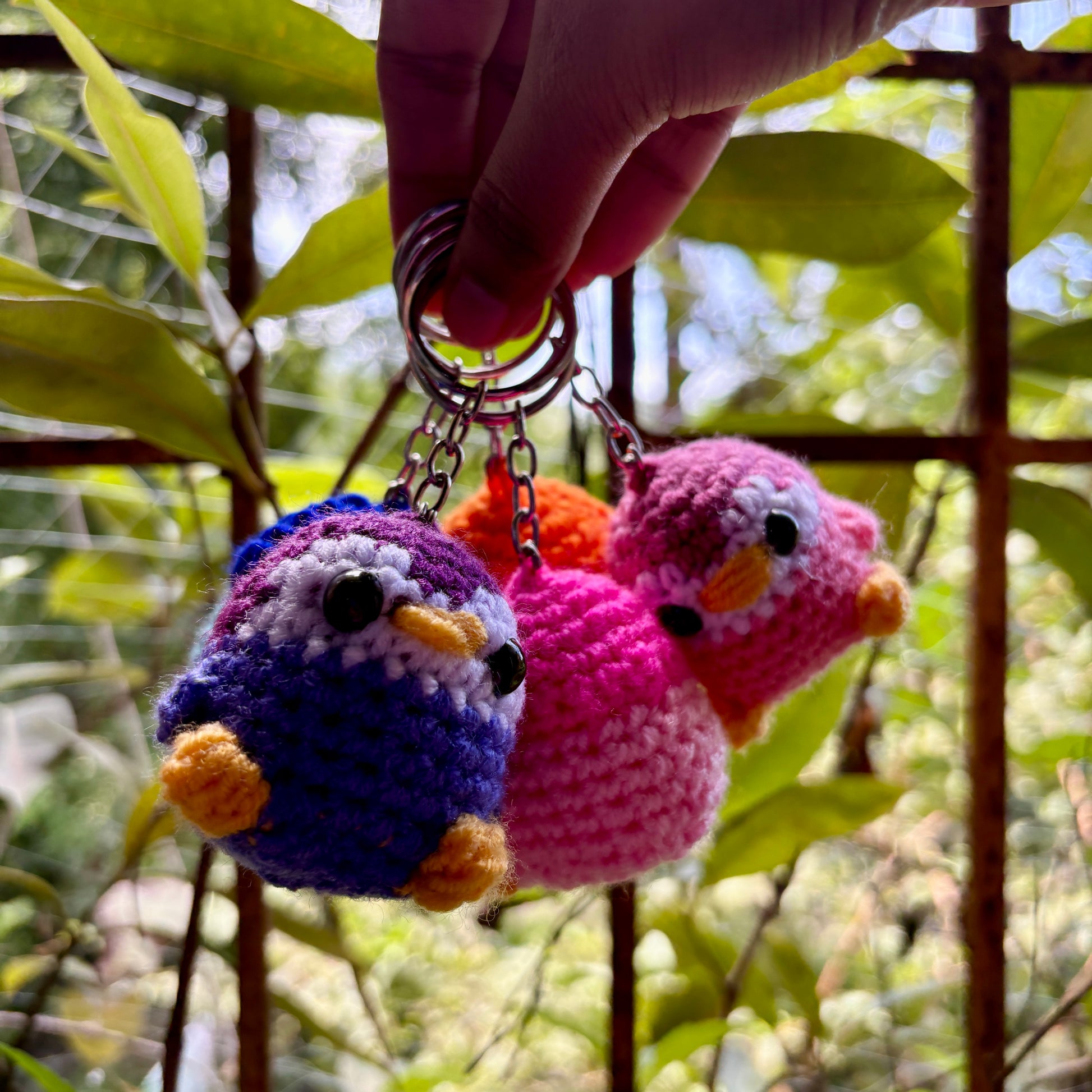 Three colorful crochet bird keychains hanging from a keychain, with a blurred natural background.