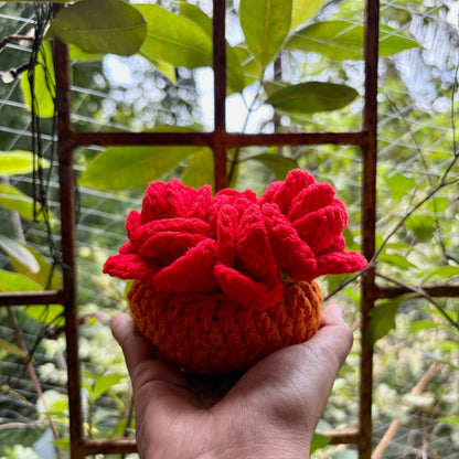Hand holding a crochet red coaster set of 3 with orange pot against a blurred green outdoor background