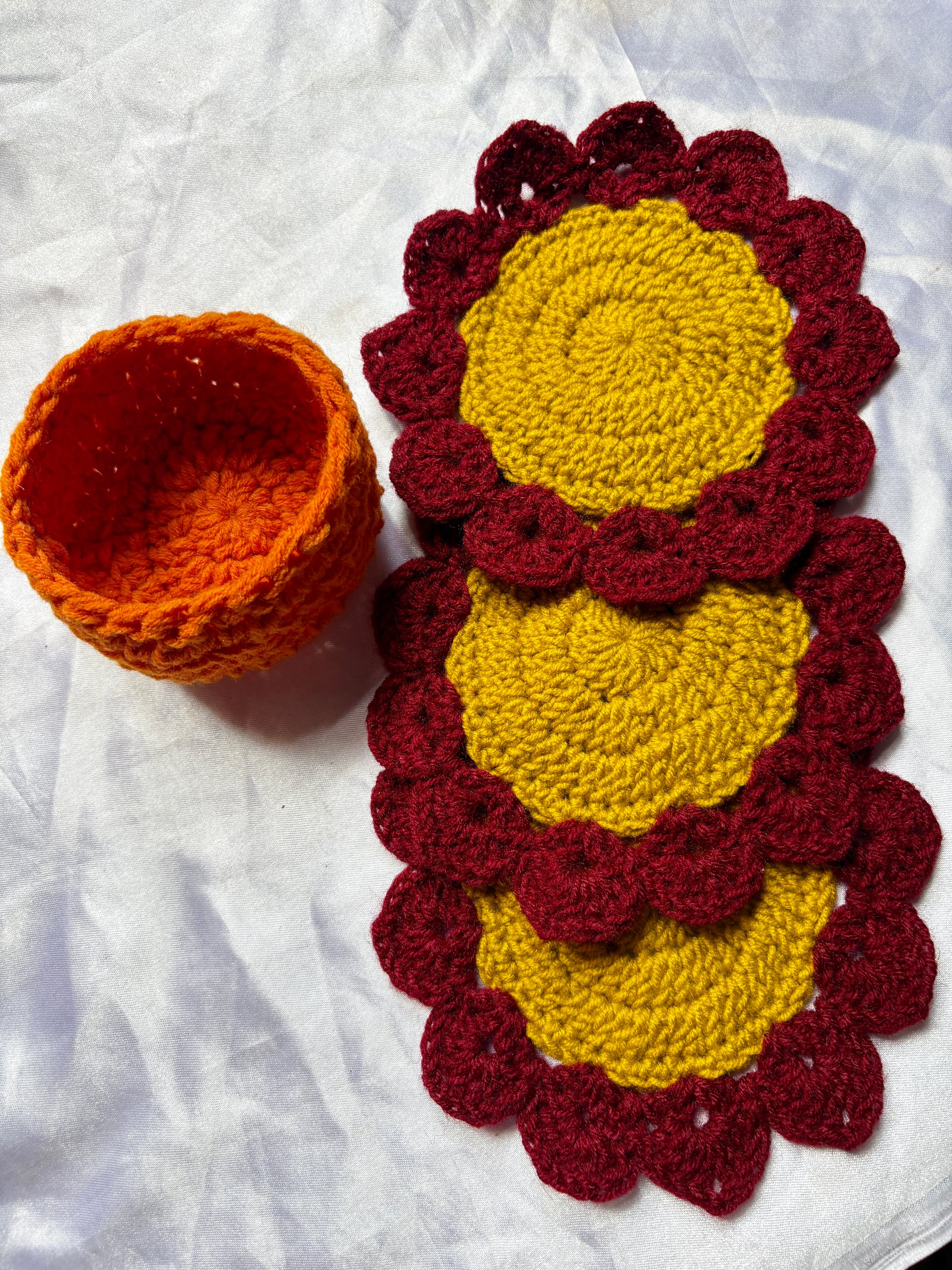 Crocheted coasters with yellow centers and maroon outer layers, alongside an orange bowl on a white fabric background.