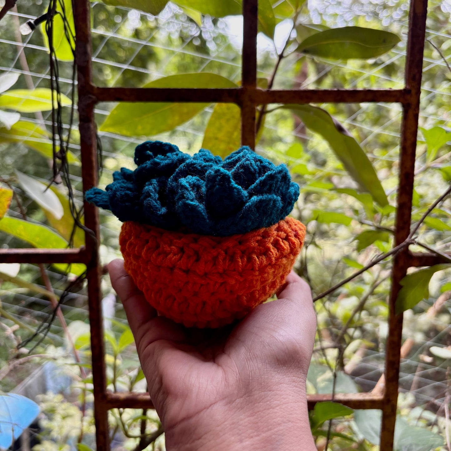 Hand holding a crochet Teal coaster set of 3 with orange pot against a blurred green outdoor background
