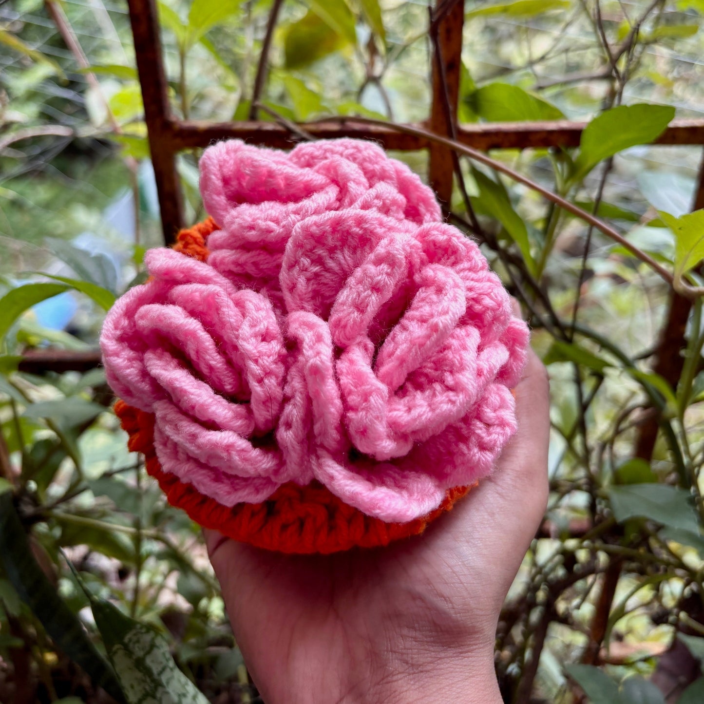 Hand holding a crochet pink coaster set of 3 with orange pot against a blurred green outdoor background