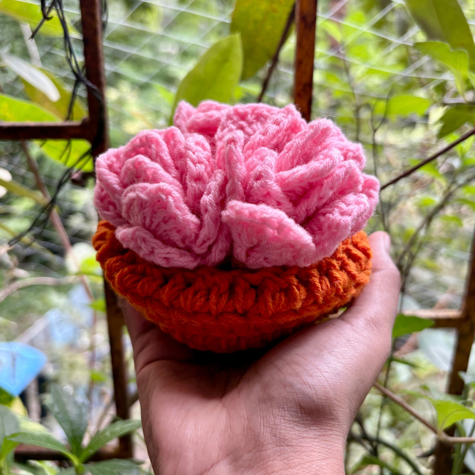 Hand holding a crochet pink coaster set of 3 with orange pot against a blurred green outdoor background