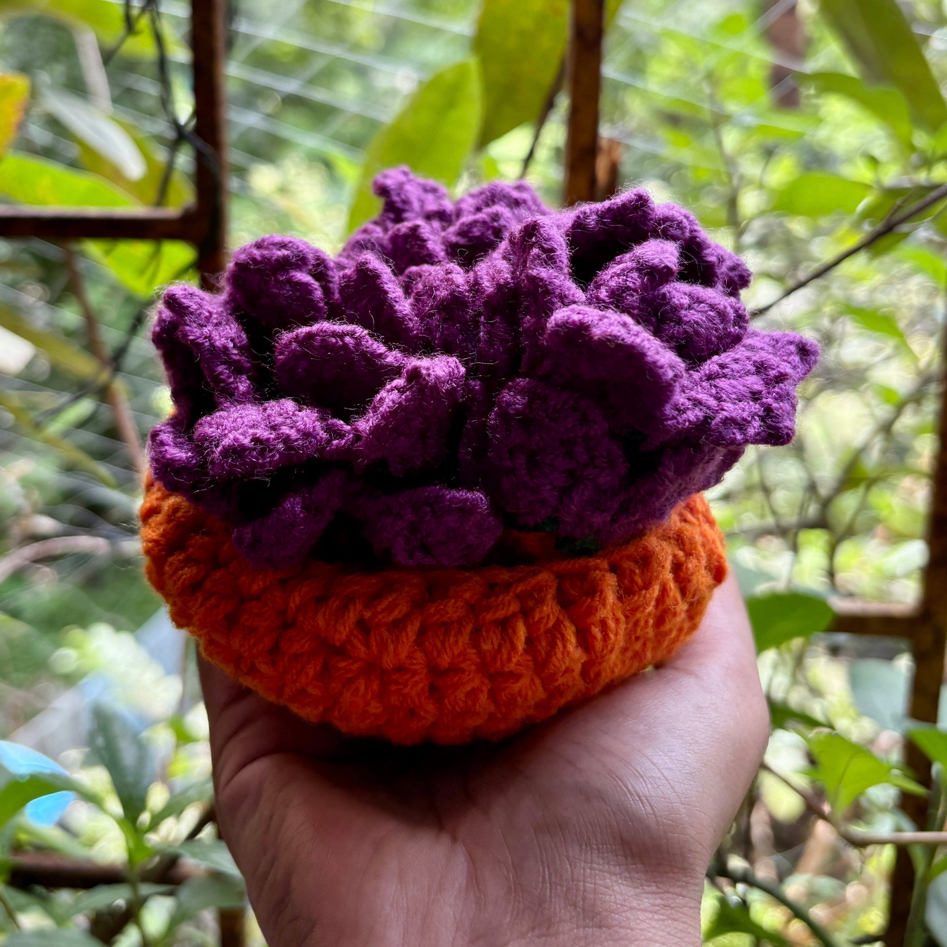 Hand holding a crochet purple coaster set of 3 with orange pot against a blurred green outdoor background