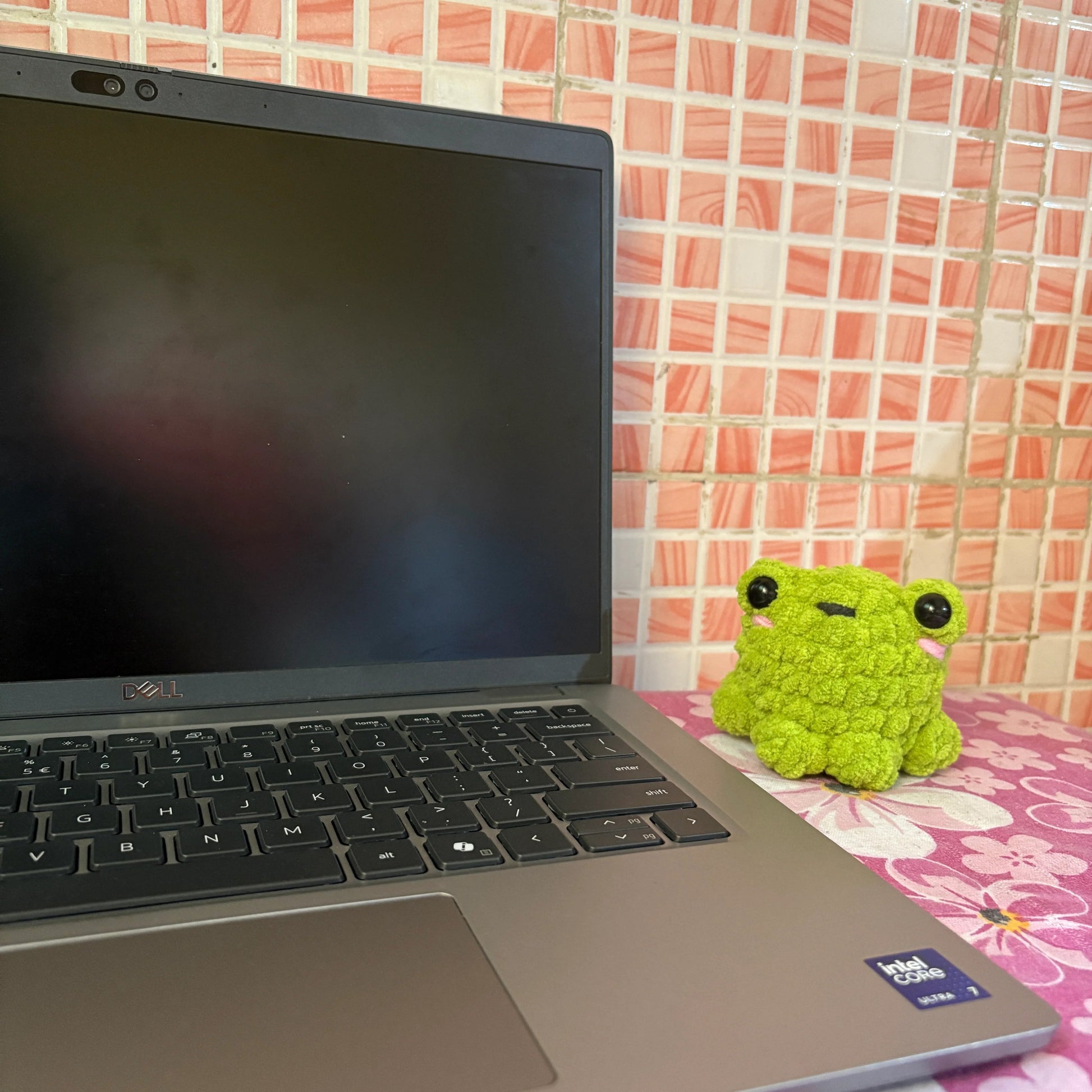 Laptop on a pink floral surface with a small green frog plush toy next to it against a tiled wall.